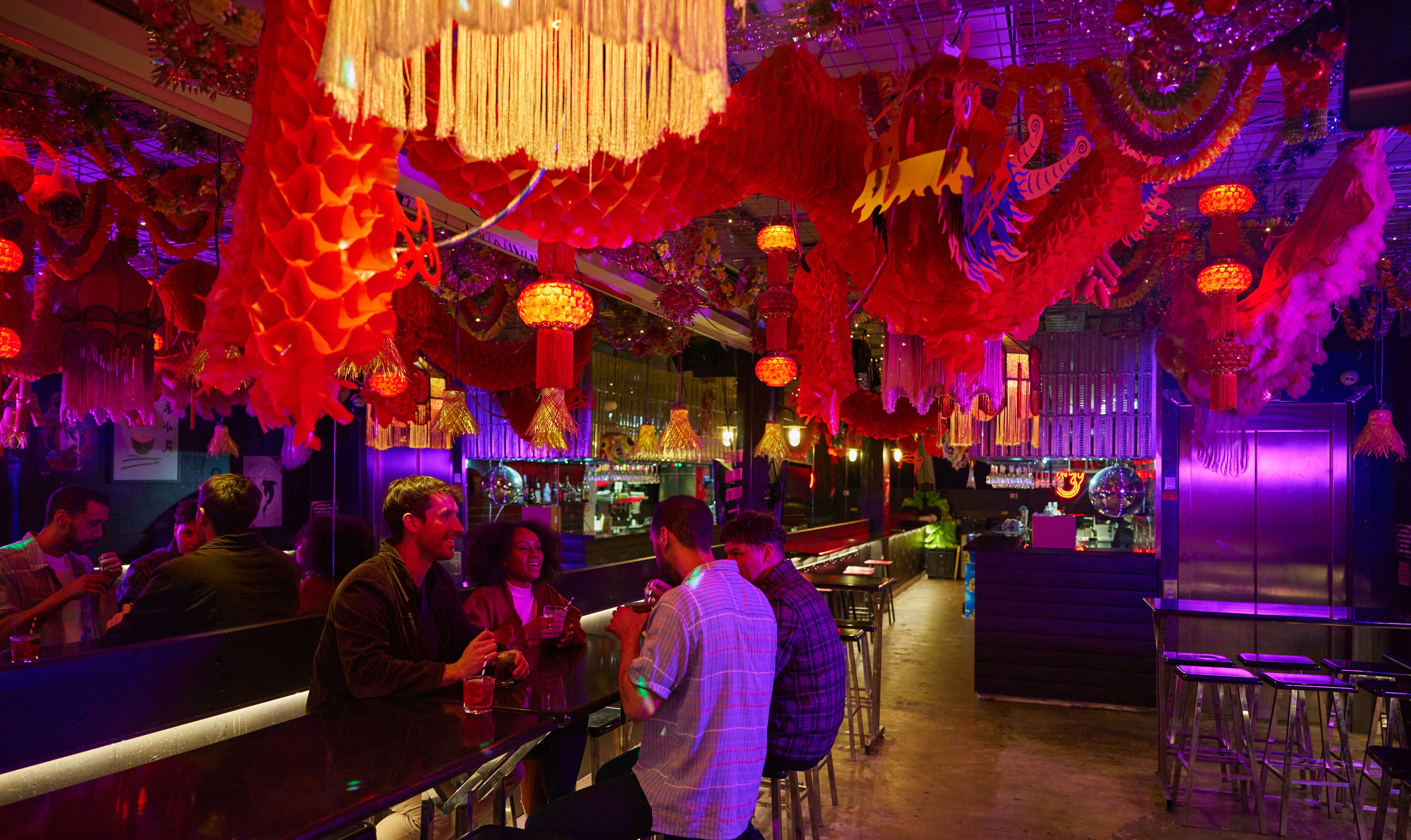 Four people drinking in a neon-lit bar with red Chinese decor hanging from the ceiling.
