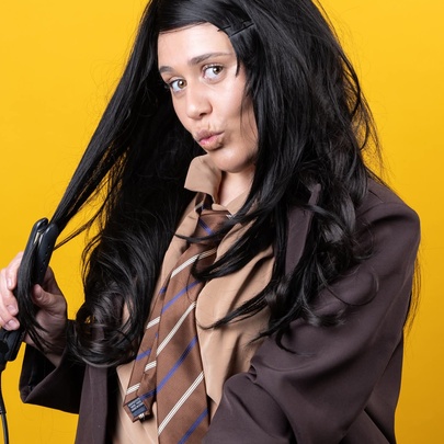 A woman dressed in a brown-toned school uniform straightening her long brown hair with a hair straightener.