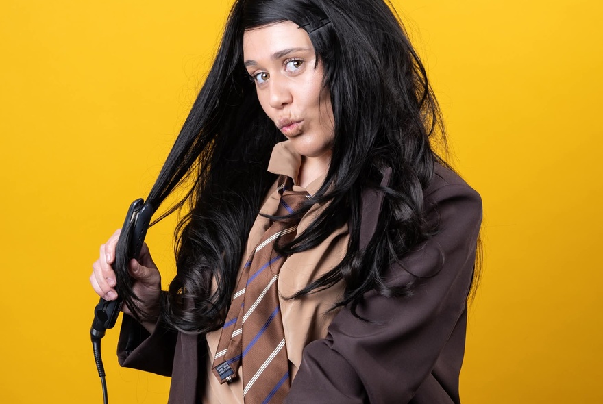 A woman dressed in a brown-toned school uniform straightening her long brown hair with a hair straightener.