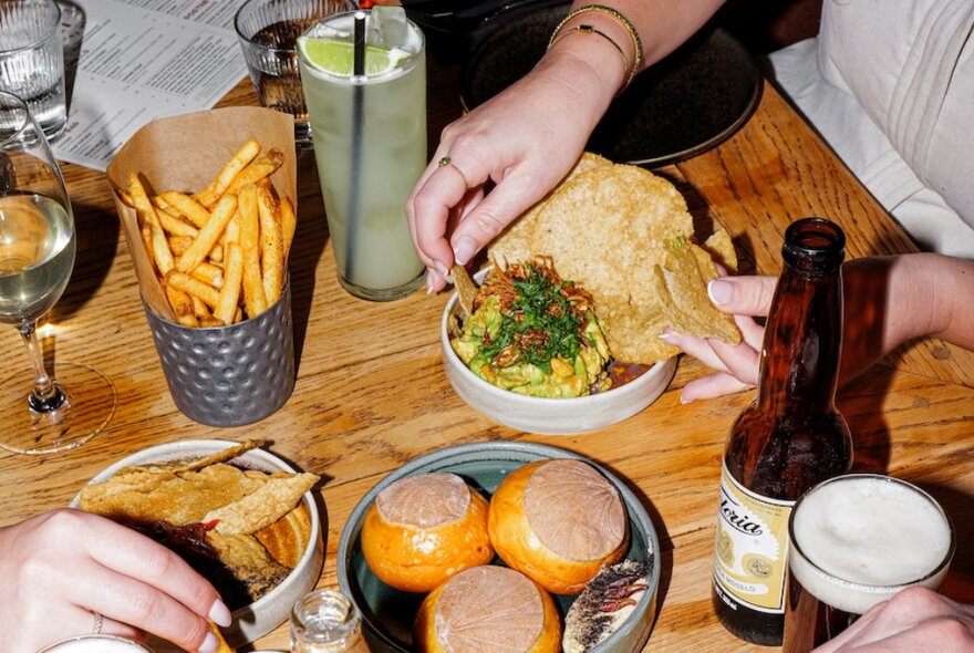 Looking at a table with hands reaching for Mexican food, chips, beer and cocktails. 