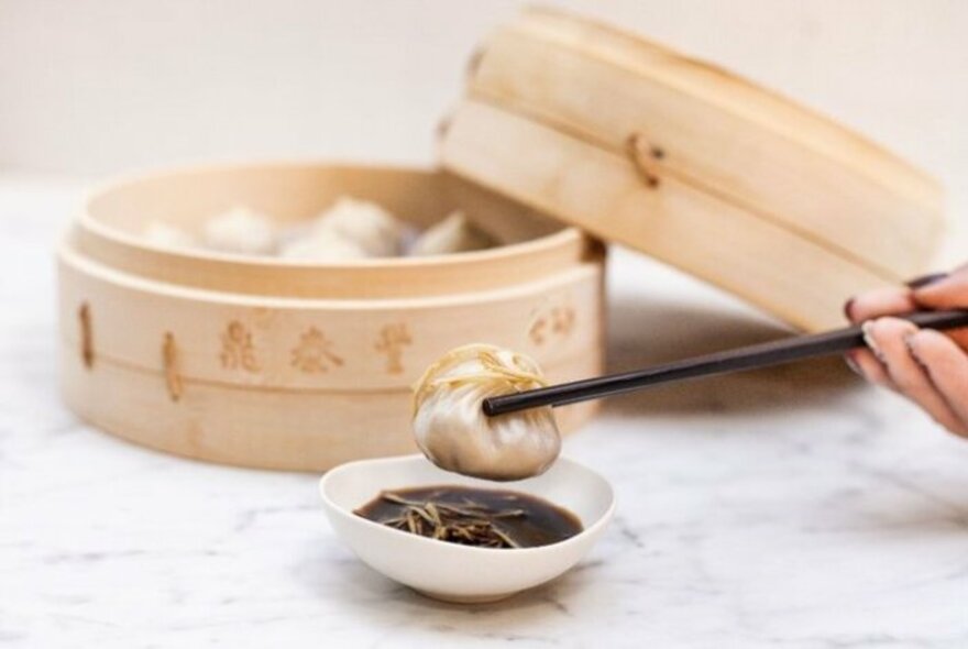 Bamboo container and person using chopsticks and dipping a dumpling in sauce.