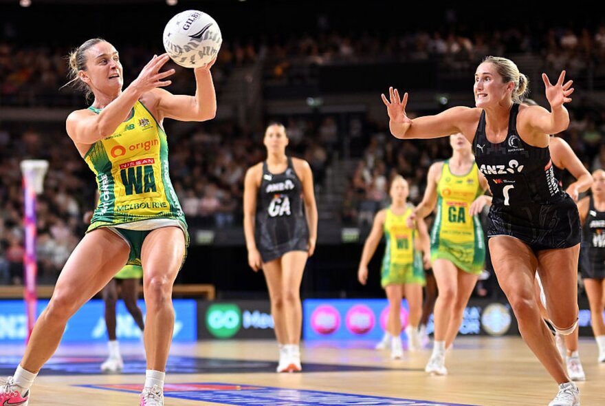 A netball player in a green and gold uniform catches the ball while a defender in a black uniform lunges toward her on an indoor court, blurred players and seated fans in the background.