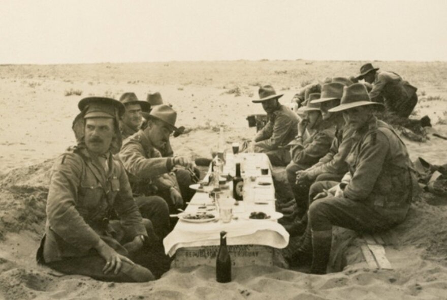 A group of soldiers eating a meal at a makeshift table dug out of sand, with a tablecloth, proper plates and two bottles of beer.