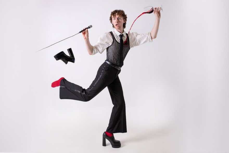 Performer Tomas Clifford posing against a white background with a bemused expression on his face, kicking his back leg out behind him and losing his chunky platform shoe in the process, holding a microphone in one hand and spilling a glass of red wine over his shirt with his other hand.