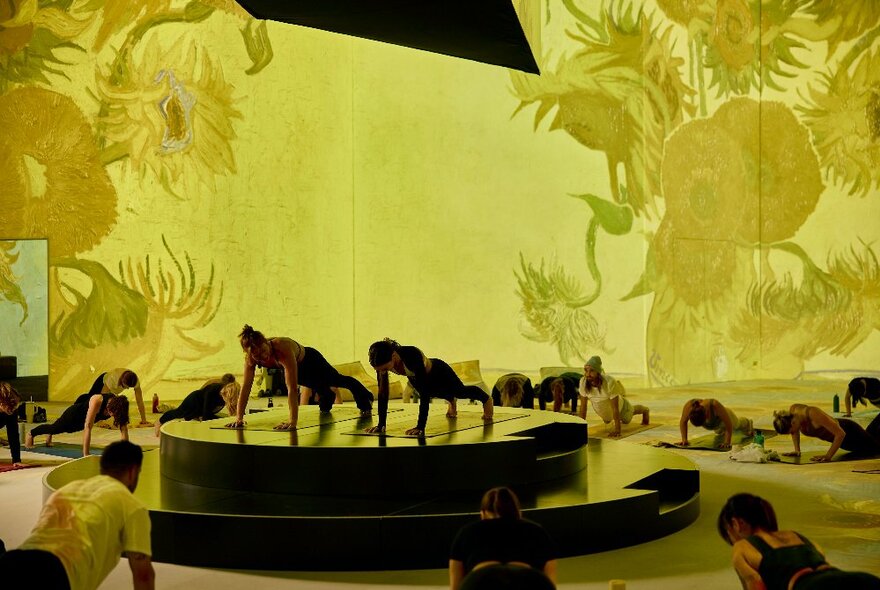 Wellness class exercising in a circle around people doing pushups on a central dais with a backdrop of Van Gogh's sunflower imagery.