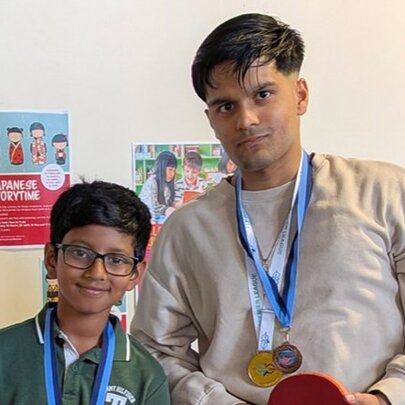 A child and an adult with sporting medals around their necks, one of them holding a table tennis paddle, standing in a room.