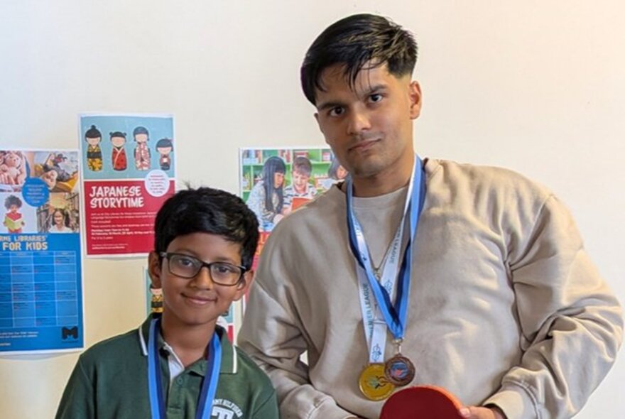 A child and an adult with sporting medals around their necks, one of them holding a table tennis paddle, standing in a room.