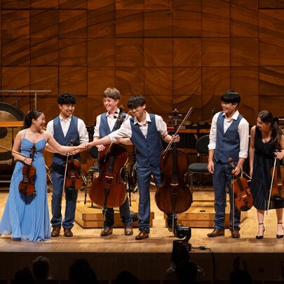 A string ensemble of six young musicians bowing on stage in a recital hall.
