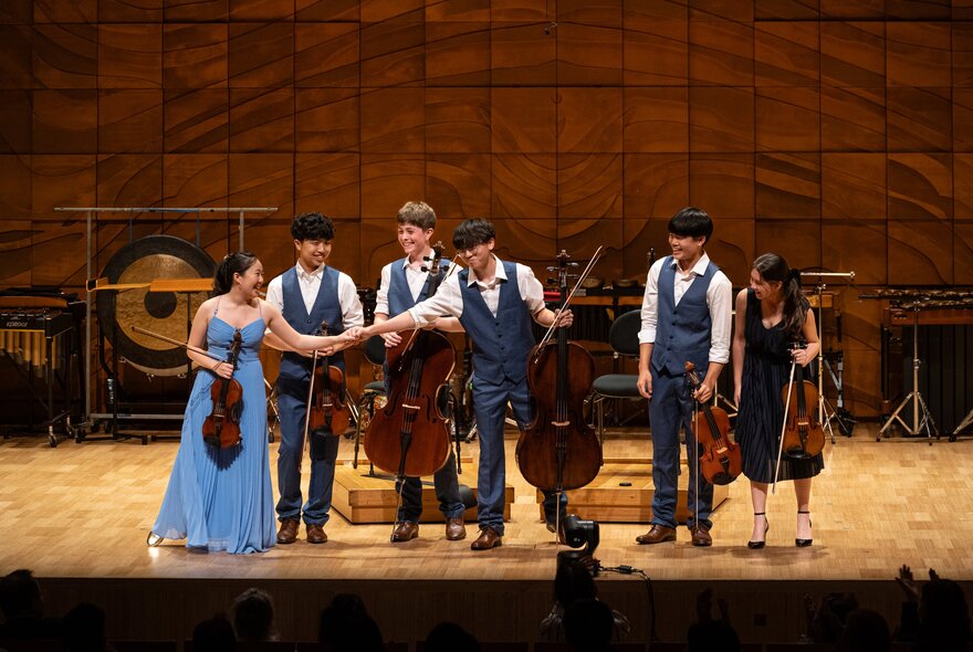 A string ensemble of six young musicians bowing on stage in a recital hall.