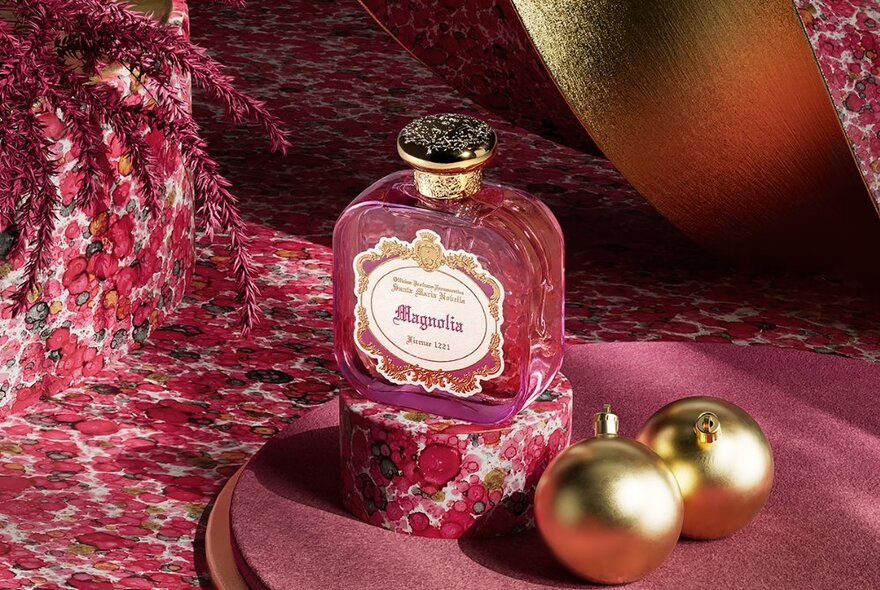 Bottle of pink perfume and gold Christmas baubles on a pink floral background.