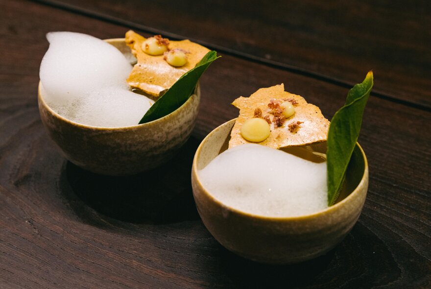 Two foamy cocktails presented in small earthenware bowls, each with a bay leaf and crisp biscotti as garnish.
