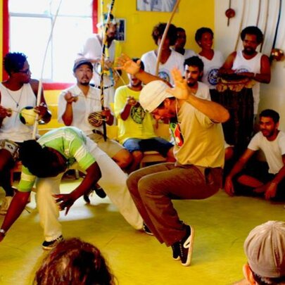 People participating in a Capoeira Angola class, with two people dancing and moving in the centre of a seated group of onlookers. 
