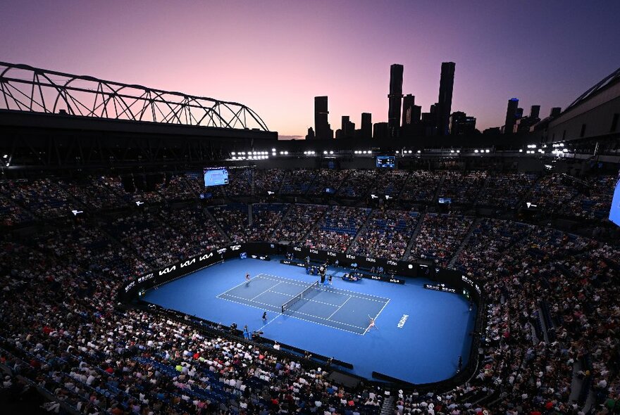 Melbourne Park arena at twilight, with city buildings against purple night sky and main court lit up.