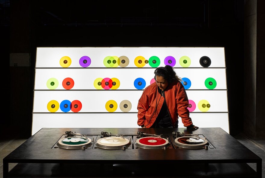 Interactive installation at The Vinyl Factory: Reverb exhibition shows a person wearing headphones standing behind a table with four turntables, in front of a brightly lit display wall featuring rows of small, colorful vinyl records.