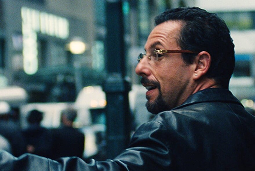 A man wearing glasses in profile looking in charge and serious in a room with people out of focus in the background. 