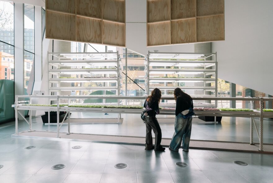 Two people stand in the bright, modern interior of the Science Gallery space, looking down at something, in the background are large windows and rows of white shelves.
