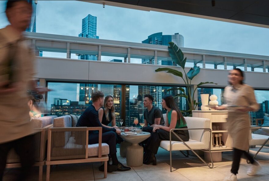 A group of people enjoying a drink at a rooftop bar with the city skyline at dusk behind them.