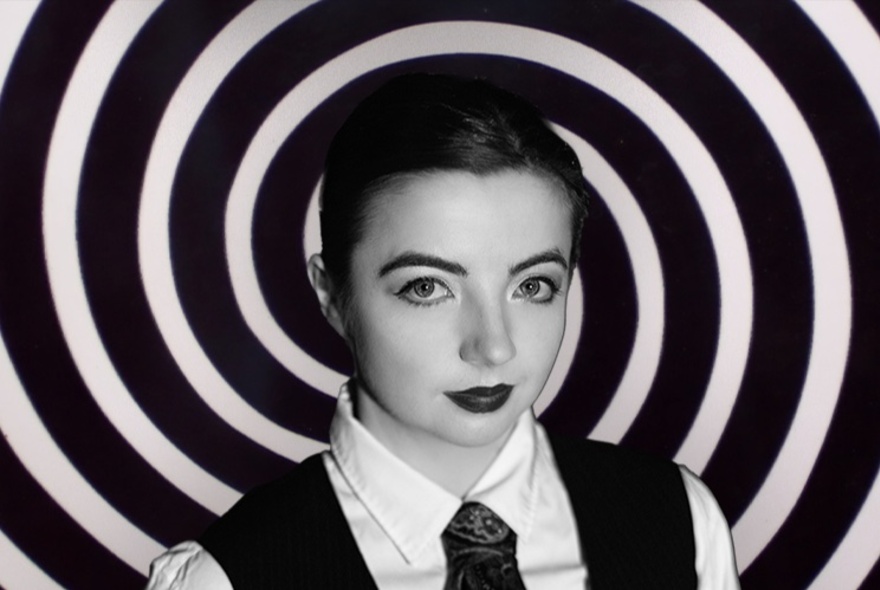 Woman wearing a tie and waistcoat with short dark hair and lipstick, circular pattern on the wall behind her.