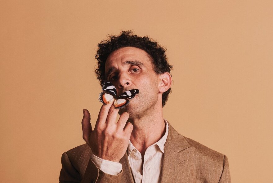 Actor wearing a tan coloured suit jacket, with a butterfly resting on his fingers held up near his face and covering his mouth.