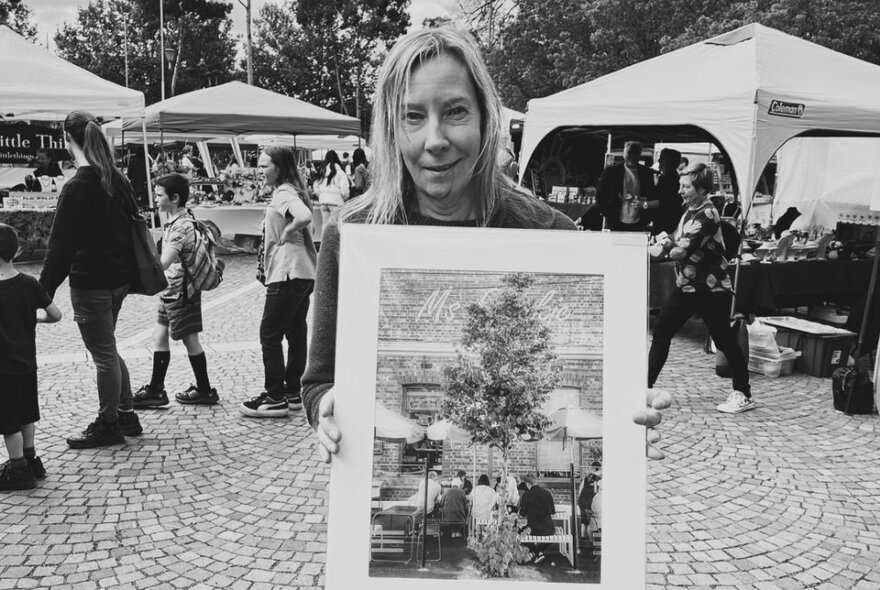 Woman holding a large artwork in a paved market area with people queueing behind her next to tents.