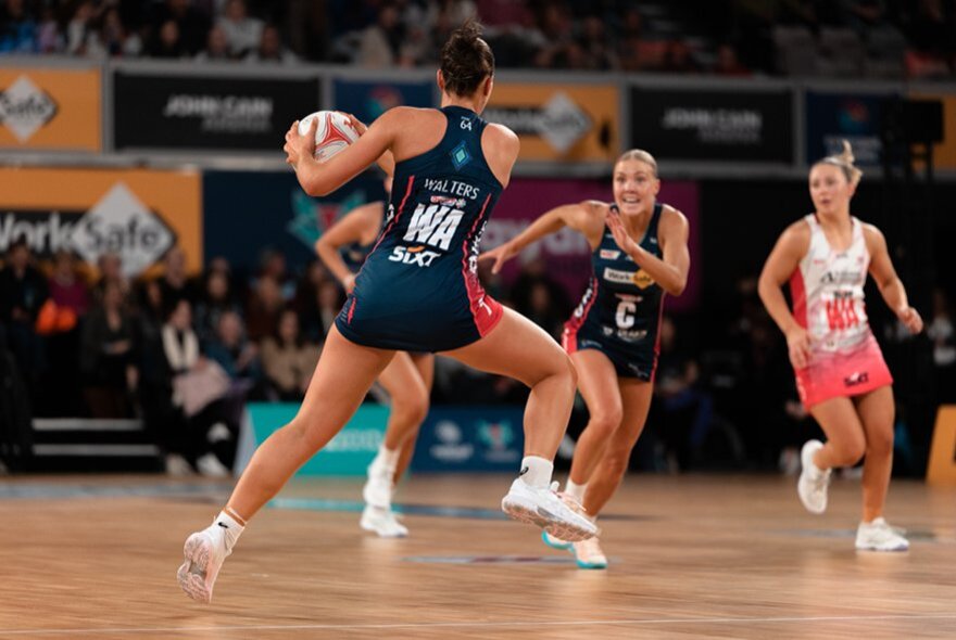 An action shot of a netballer on court with the ball, other netballers running.