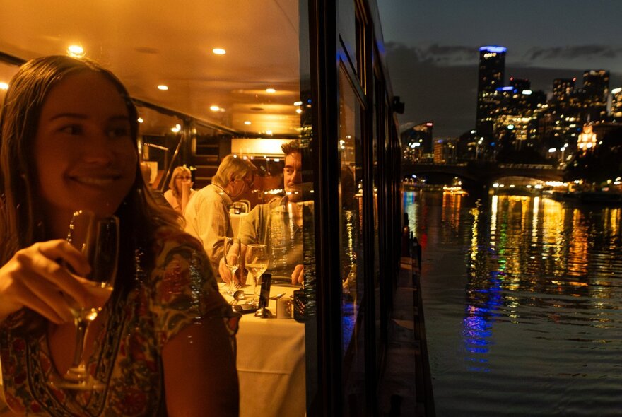 People dining on board a boat, with a view of the Yarra River and the city skyline in the background.
