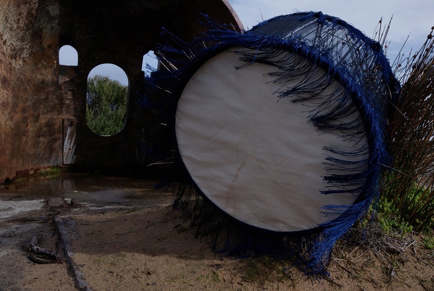 An outside drum with a blue fringe, sitting on a sandy area with a building structure behind it.