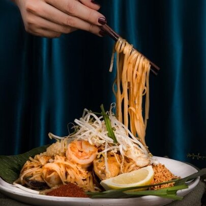 Hand using chopsticks to lift noodles from a  plate of Pad Thai featuring rice noodles, prawns, bean sprouts, crushed peanuts, and a fresh wedge of lemon.