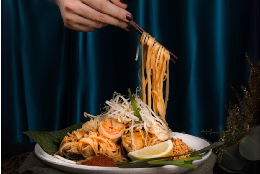 Hand using chopsticks to lift noodles from a  plate of Pad Thai featuring rice noodles, prawns, bean sprouts, crushed peanuts, and a fresh wedge of lemon.