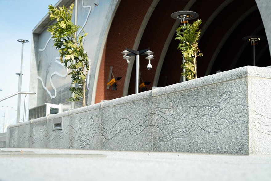 Indigenous artwork carved into the stone used in low fencing along a pathway; entrance to a large archway in the background.