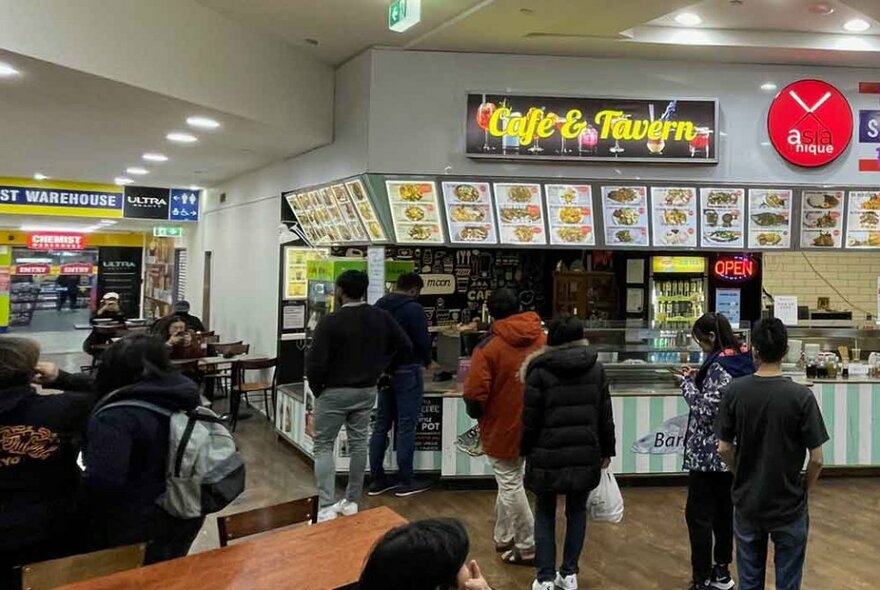 View into part of the retail food court at the Paramount Centre with people lining up at a food outlet and some eating at tables and chairs.