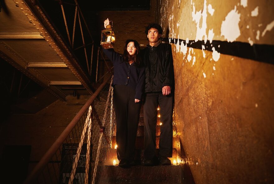 Two people descending dimly-lit narrow stairs in an old jail with mottled walls.