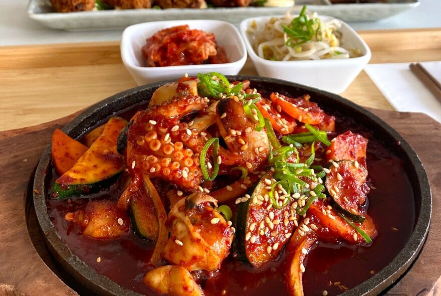 Korean spicy grilled octopus dish served on an iron cooking dish on a wooden tray, with two small white bowls with kimchi and bean shoots.