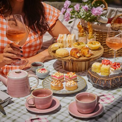 A checked tablecloth with a High Tea spread on top with pink teacups and teapot and plates of cakes and sandwiches. 
