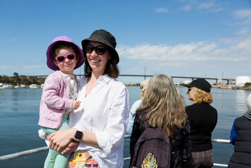 A woman holding a child on her hip outside on a boat in the sun. 