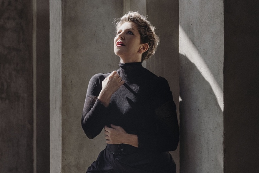 Singer Joyce DiDonato, wearing all black and posed against a grey wall, her face looking upwards and one of her hands raised near her throat.