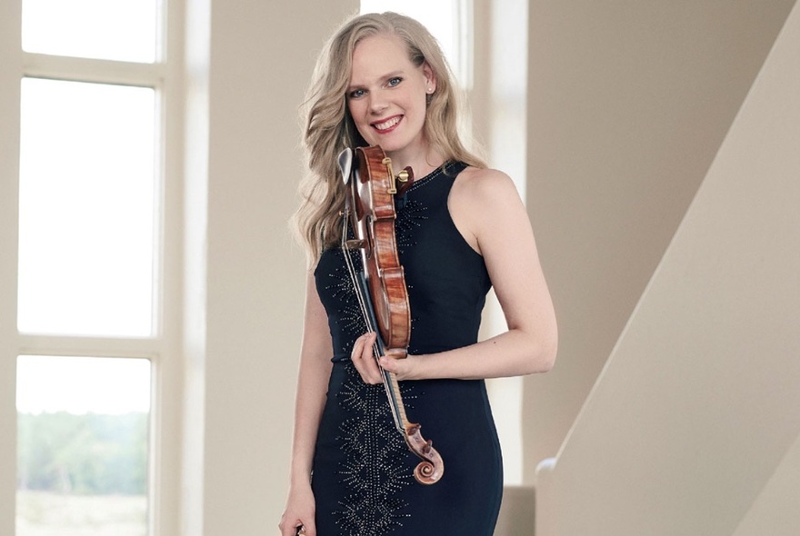 Portrait of violin virtuoso, Simone Lamsma, posing in a black dress and holding her violin, in front of a white staircase and windows.