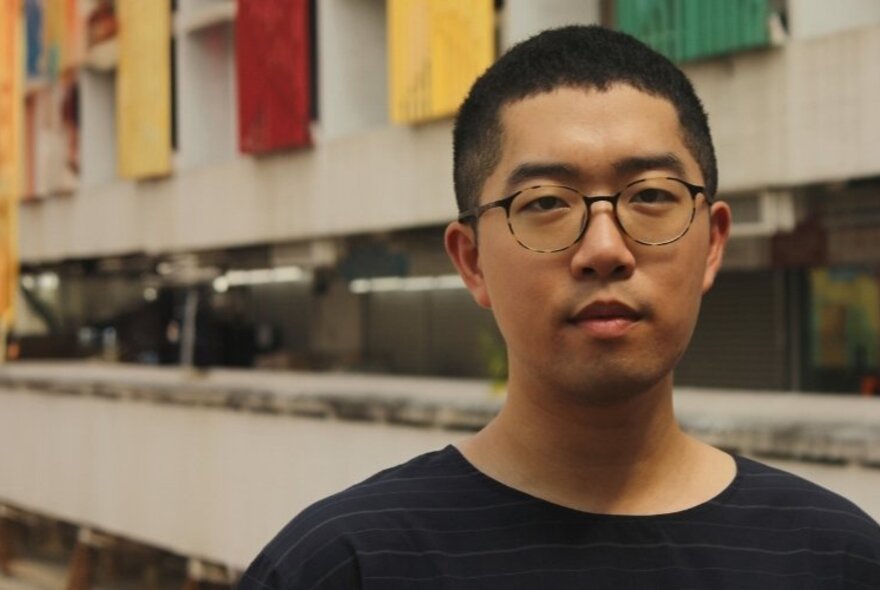 Portrait of a Singaporean man wearing round glasses, in front of a concrete building with colourful shutters over the windows.