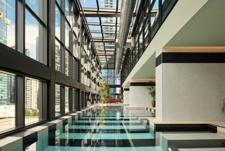 The indoor swimming pool at the Hannah St Hotel with a black and white checkered pattern at the bottom, surrounded by large, floor-to-ceiling windows and a glass ceiling, offering a view of city buildings outside. 