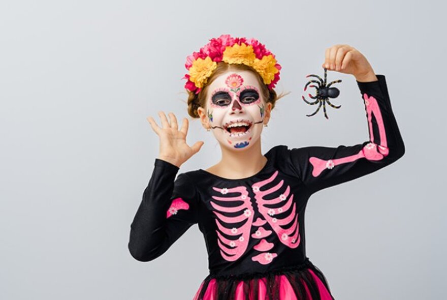 Girl in Day of the Dead costume with pink rib cage on a black background, skull makeup and floral headdress, holding a black spider.