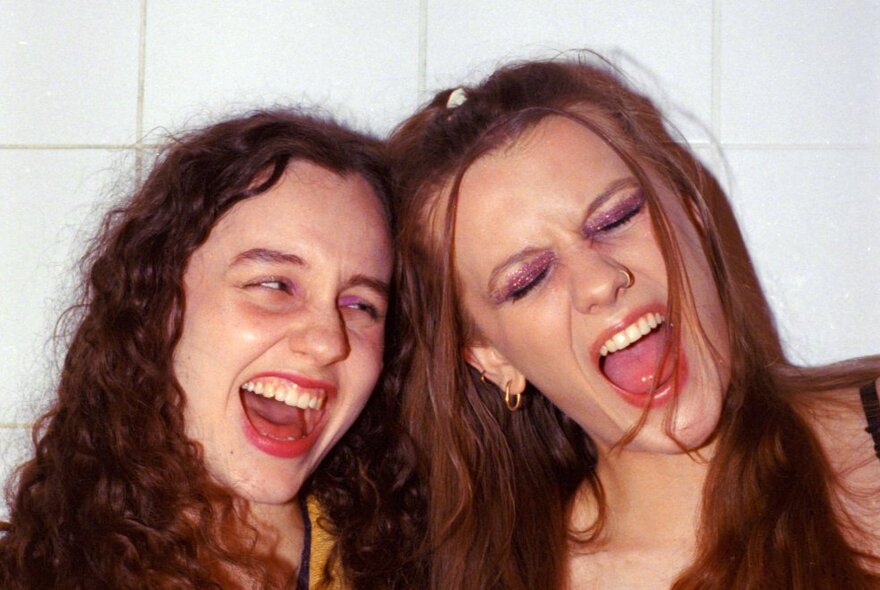 Two girls with their heads together laughing against a white tiled wall.