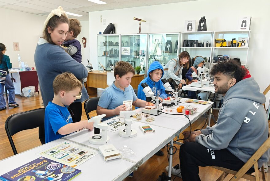 Children and adults seated at long workshop tables working with specimens and microscopes.