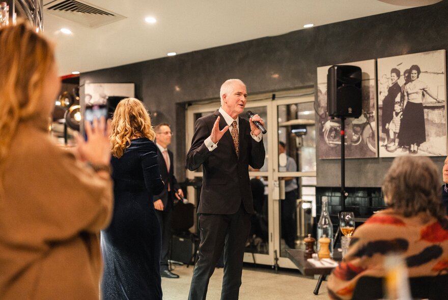 A tenor singer in a black suit singing into a microphone at an Italian restaurant; patrons watching on.