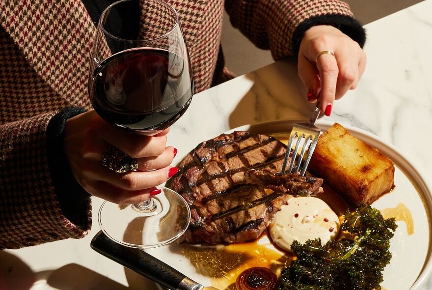 Hand holding a large glass of red wine in one hand, a fork over a large piece of meat on a plate in the other.