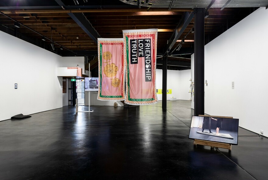 Exhibition space with polished black floors, white walls and two banner style artworks hanging from the ceiling.