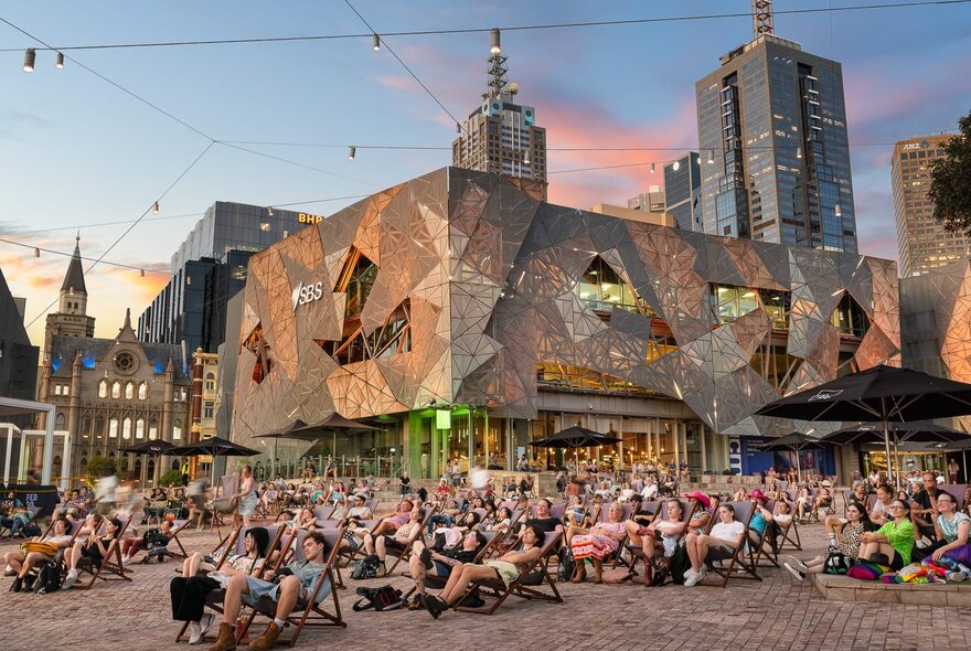 A crowd of people sitting in Fed Square's open plaza at dusk, watching a projection on Fed Square's big digital screen (out of the frame).
