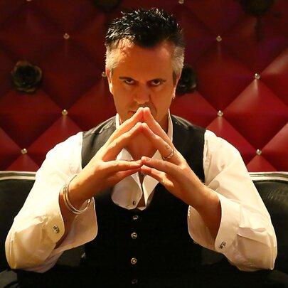 Magician Duck Cameron, a man seated on a small black couch with his hands held up in front of his face, with fingertips touching.