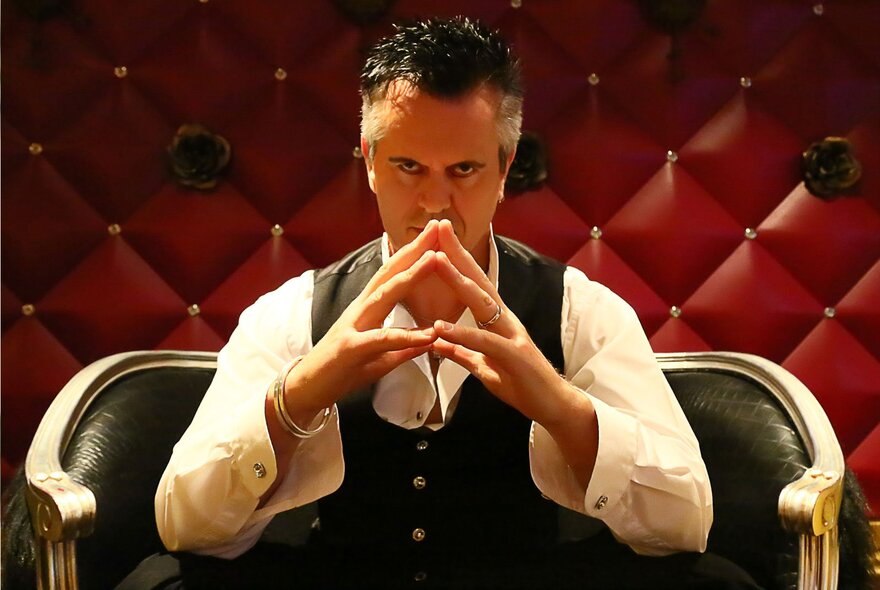 Magician Duck Cameron, a man seated on a small black couch with his hands held up in front of his face, with fingertips touching.