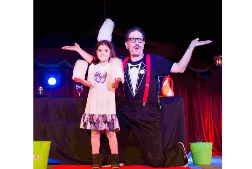 Performer and child during a magic act on stage, their arms outstretched.
