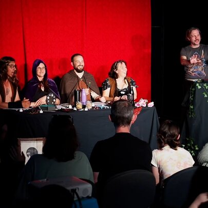 Four performers seated at a table on a stage behind a table, with a person guiding them through a Dungeons and Dragons style improvised adventure. 